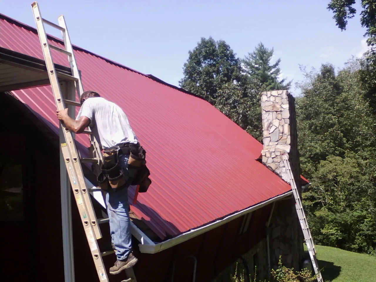 Roofer on a ladder beside a red metal roof and stone chimney — Soffit & Fascia Repair by Roofing Brothers in Windham